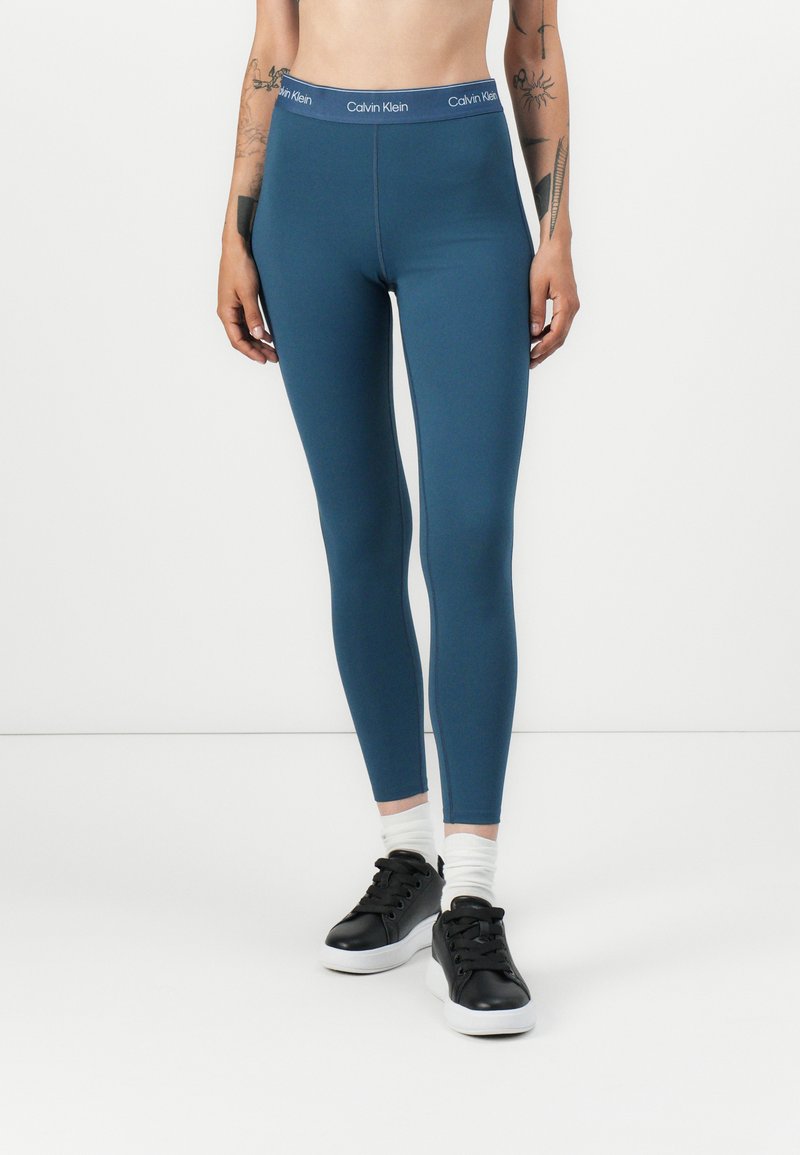 Teal tajice visokog struka s glatkom teksturom, sa pojasom s Calvin Klein logotipom i krojem koji se čvrsto prilagođava tijelu. Uparene s crnim tenisicama.