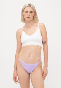 Bralette branco com alças finas e decote V profundo, combinado com uma thong de corte baixo em lavanda com uma faixa do logótipo. Textura suave, design minimalista.
