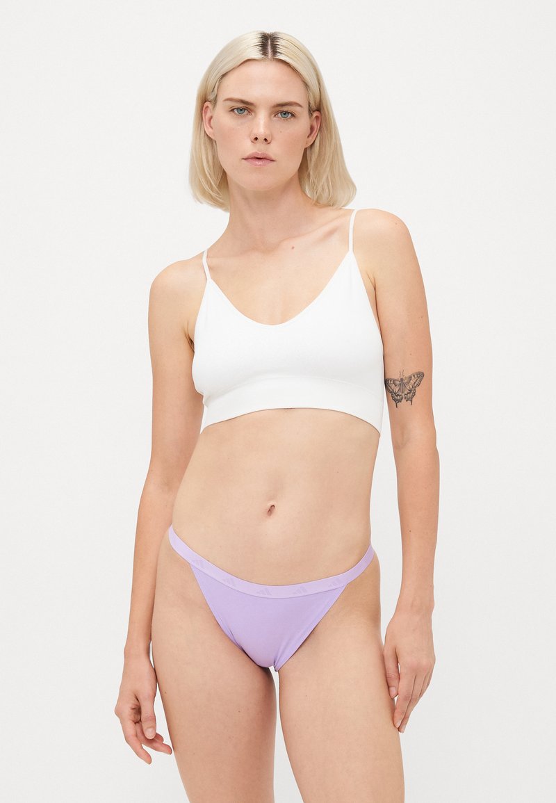 Bralette branco com alças finas e decote V profundo, combinado com uma thong de corte baixo em lavanda com uma faixa do logótipo. Textura suave, design minimalista.