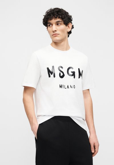 Homme aux cheveux bouclés portant un t-shirt blanc graphique MSGN Milano et un pantalon noir, debout avec une main dans la poche, regardant sur le côté.