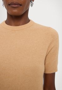 Haut à manches courtes en cachemire beige doux avec col rond. Présente une texture tricotée fine et des bords côtelés. Détail des boucles d'oreilles visible.