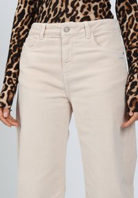 Beige Cordhosen mit vertikalen Rippungen, fünf Taschen und einem Knopfverschluss. Kombiniert mit einem figurbetonten Langarmshirt mit Leopardenmuster.