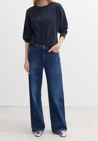 Marinblå sweatshirt med sänkta axlar, kombinerad med blå jeans med hög midja och vida ben. Modellen bär metallicsömlösa silverskor.