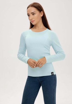 Frau mit langen braunen Haaren, die ein hellblaues Langarmshirt und dunkelblaue Jeans trägt, steht vor einem einfarbigen Hintergrund.