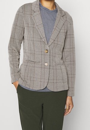 Frau trägt ein beige-schwarz kariertes Sakko mit zwei Knöpfen über einem grauen Hemd mit Rüschenkragen und dunkelgrünen Hosen.
