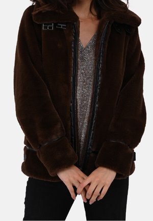 Kobieta ubrana w ciemnobrązową kurtkę z sztucznego futra z klamrami na srebrnym błyszczącym topie z dekoltem w serek, ręce splecione z przodu, widoczne czarne spodnie.