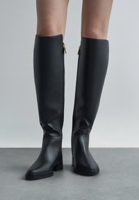 Bottes en caoutchouc noires jusqu'aux genoux avec une texture lisse, orteils carrés, talons blocs et détails de fermeture éclair dorée à l'arrière.