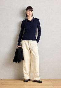 Polo à manches longues bleu marine avec détail de boutons, associé à un pantalon large crème. Le mannequin tient un pull noir dans un cadre minimaliste.