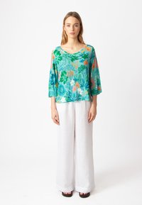 Blusa a maniche corte con fantasia floreale in turchese, che presenta colori vivaci. Abbinata a pantaloni in lino bianchi larghi e sandali neri.