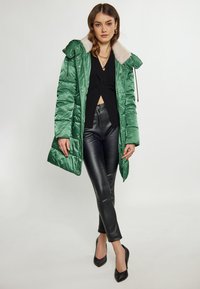 Jachetă parka verde căptușită cu guler de blană, purtată peste un top negru, asociată cu pantaloni negri mulați și tocuri negre. Material neted, lucios.