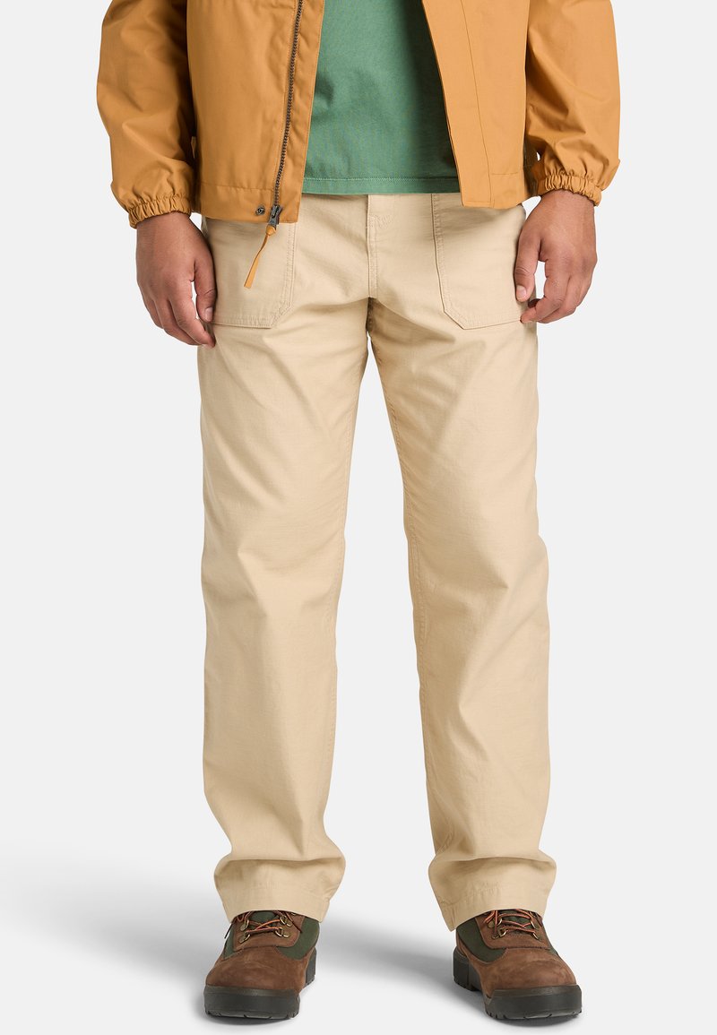 Beige Baumwollhosen mit geradem Bein, ausgestattet mit Taschen vorne, verstärkten Nähten und einer lockeren Passform, kombiniert mit braunen Wanderschuhen.