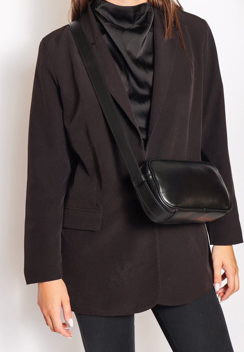 Blazer nero su una top in satin nero, abbinato a una piccola borsa a tracolla nera. La borsa presenta una finitura elegante e liscia e una tracolla regolabile.