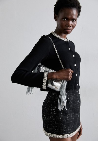 Crna skraćena kardigan sa srebrnim dugmadi, ukrašenim manžetama, i uzorčana crna mini suknja sa srebrnim akcentima. Drži srebrnu torbu sa resicama.