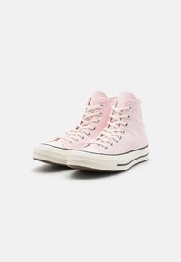 Høye sneakers i myk rosa kanvas med hvite snørebånd, en gummitåhette og en beige såle med svarte aksentstriper.