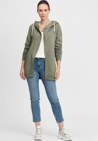 Cardigan verde con cappuccio, dotato di zip completa, texture a maglia e lacci color crema, indossato sopra una maglietta bianca e jeans blu, abbinato a sneakers.