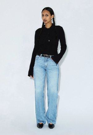 Frau mit geflochtenem Haar, die ein schwarzes Langarmshirt, hellblaue Jeans, einen schwarzen Gürtel und schwarze spitze Schuhe trägt, steht vor einem schlichten weißen Hintergrund.