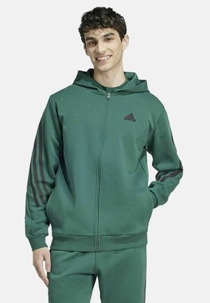 Grüne, mit Reißverschluss und Kapuze ausgestattete Sweatjacke mit schwarzen Streifen an den Ärmeln. Verfügt über ein Logo auf der Brust, ein weiches Material und ein lässiges Design.