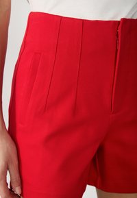 Červené šortky z hladkej látky, vybavené bočnými vreckami, vysokým pásom a predným prúžkovaným detailom. Zapínanie na zips na boku.