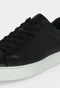 Baskets basses noires avec lacets noirs et semelle en caoutchouc blanche, montrant les détails latéraux et avant sur fond gris.
