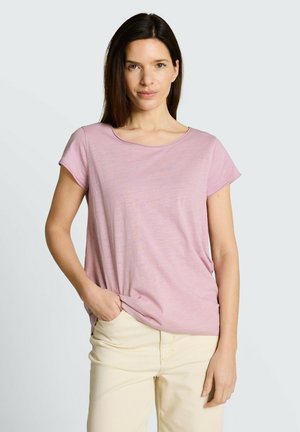 Vrouw met steil bruin haar, gekleed in een lichtpaarse top met korte mouwen en beige broek, staand met één hand in de zak tegen een effen achtergrond.