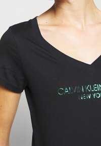 Black V-neck t-shirt with short sleeves, featuring shiny green "Calvin Klein New York" text on the chest, worn by a person.