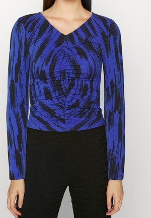 Frau trägt ein langärmeliges Oberteil in Blau und Schwarz mit abstraktem Pinselstrichmuster, kombiniert mit texturierten schwarzen Hosen, steht vor weißem Hintergrund.