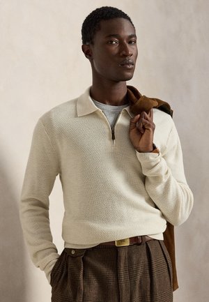 Homme portant un polo à manches longues texturé crème et un pantalon marron à carreaux tient une veste marron sur son épaule sur un fond beige.