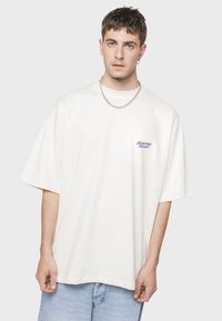 Camiseta oversized color crema hecha de algodón, con un pequeño logo púrpura de "Kaotiko Resort" en el lado izquierdo. Combinada con pantalones cortos de mezclilla.