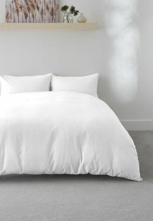 Weißes Bett mit zwei Kissen und einer weißen Bettdecke auf einem grauen Teppich, mit einem Holzregal, das Vasen und ein Gemälde darüber hält.