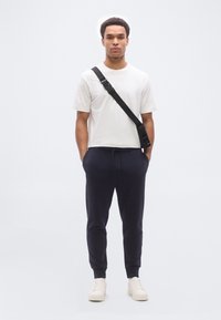 Άνδρας που φοράει λευκό κοντομάνικο, μαύρη τσάντα χιαστί, σκουρόχρωμο παντελόνι φόρμας και λευκά αθλητικά παπούτσια, στέκεται με τα χέρια στις τσέπες μπροστά σε απλό φόντο.