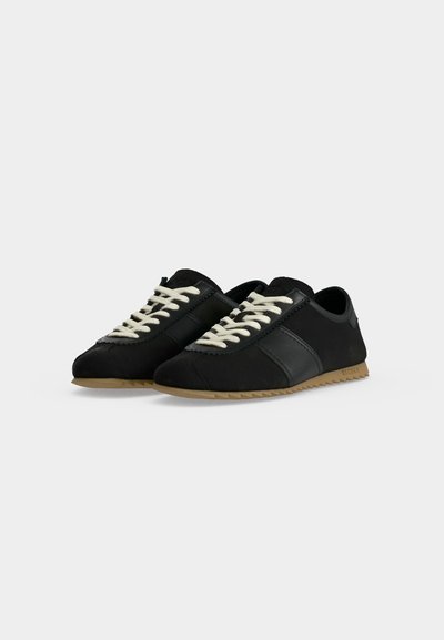 Μαύρα sneakers χαμηλού προφίλ με λευκά κορδόνια, λεία πλαϊνά πάνελ και καφέ ραβδωτές σόλες, τοποθετημένα σε απλό ανοιχτό φόντο.