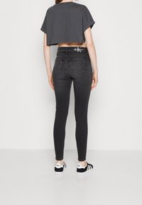 Vaqueros pitillo negros de talle alto, con bolsillos traseros y detalles de logo en la cinturilla, combinados con un top corto gris y zapatillas negras.