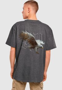 Grå bomulds T-shirt med et stort ørnegrafik trykt på ryggen, med detaljerede fjer og et fedt skriftdesign i baggrunden.