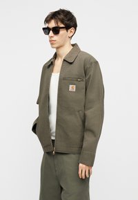 Mladý muž s okuliarmi so vzorom korytnačiny, v olivovo zelenej bunde so zipsovou náprsnou kapsou, bielom tielku a zodpovedajúcich nohaviciach, stojaci na bielom pozadí.