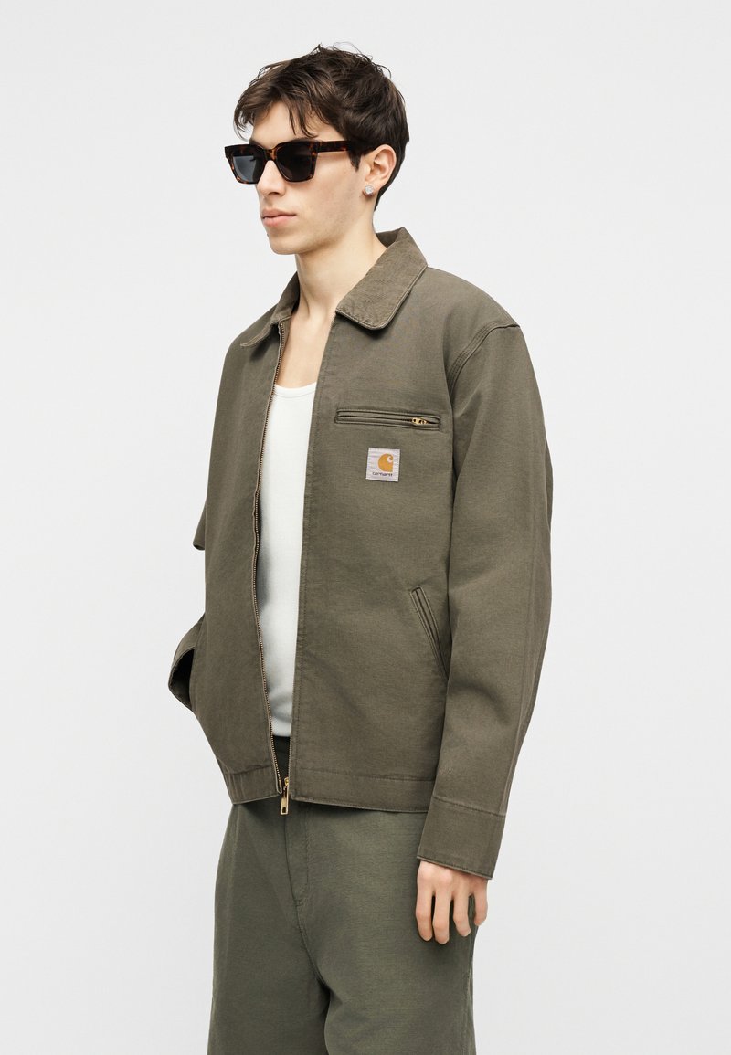 Giovane uomo con occhiali da sole tartarugati, giacca verde oliva con tasca sul petto con cerniera, canottiera bianca e pantaloni abbinati, in piedi davanti a uno sfondo bianco.