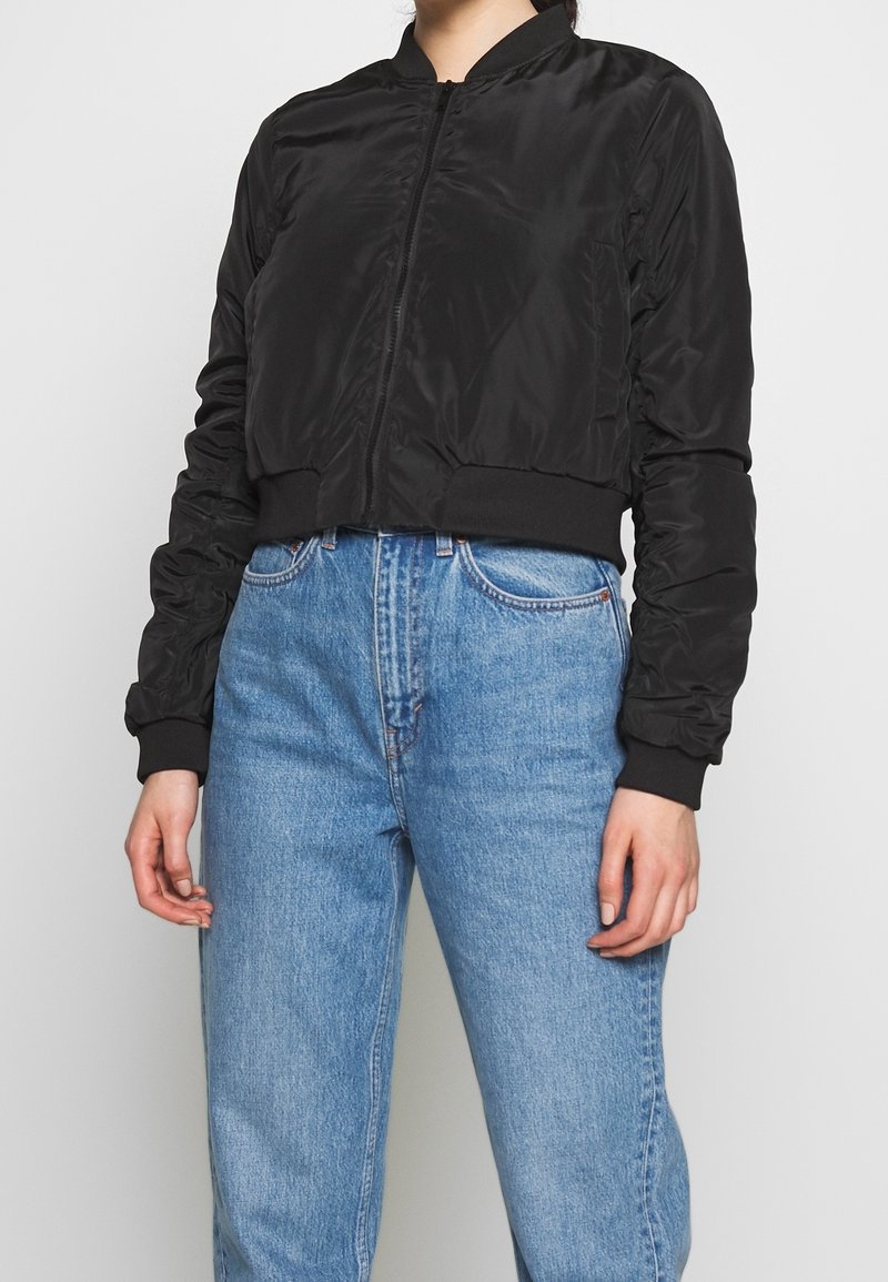 Černá zkrácená bombardérka s hladkým povrchem, žebrovanými manžetami a pasem. Spárovaná se světle modrými džíny s vysokým pasem.