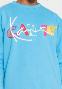 Felpa azzurra chiara con testo colorato e caratteri misti che legge "CAN'T", accompagnato da elementi grafici e un dettaglio con linee ondulate. Tessuto in cotone morbido.