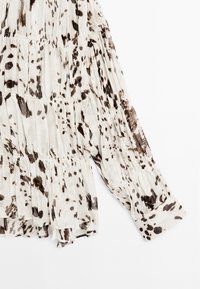 Blouse blanche avec imprimé animalier marron, présentant une texture plissée, des manches longues et un ourlet froncé. Tissu léger et poignets structurés.