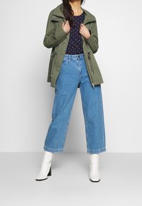 Světle olivově zelená lehká bunda s kapucí, v kombinaci s námořnickým topem s malými růžovými vzory a modrými širokými džínami s vysokým pasem. Bílé podpatkové boty.