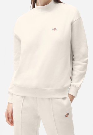 Osoba ubrana w kremową bluzę i dopasowane spodnie z małymi pomarańczowo-niebieskimi logo marki na piersi i udzie.