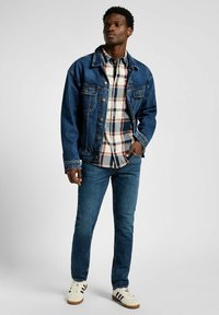 Giubbotto di jeans sopra una camicia a quadri con bottoni, jeans blu e sneaker bianche con strisce nere. Caratteristiche includono chiusure a bottone e tasche.