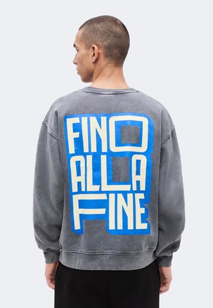 Oseba s kratkimi lasmi, obrnjena stran, oblečena v sivo jopo z velikim modro-belim napisom "FINO ALLA FINE" na hrbtu.