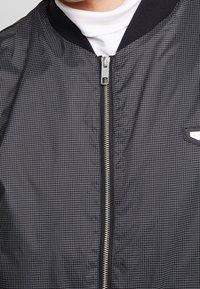 Svart bomberjacka med rutmönster, dragkedjestängning, ribbad krage och en liten logotypapplikation på framsidan. Slät texturerad tyg.