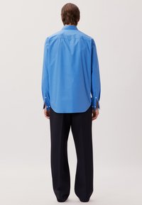 Langärmliges blaues Hemd aus Baumwolle mit abgerundetem Saum, Knopfmanschetten und einer Schulternaht hinten, kombiniert mit dunkler, lockerer Hose.