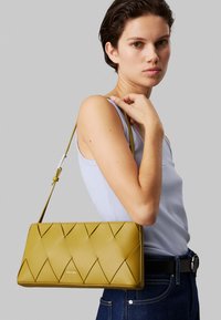 Borsa in pelle gialla senape con un motivo trapuntato e una lunga tracolla, esposta su uno sfondo grigio. Presenta un logo nella parte inferiore.