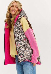Pufová vesta s leopardím vzorem, růžovou podšívkou a kapucí. Vyznačuje se kombinací materiálů a geometrickým barevným blokováním.