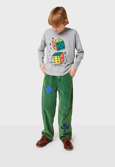 Graues Langarmhemd mit buntem Würfelmotiv, kombiniert mit grünen Cordhosen, die blaue und marineblaue Patches aufweisen. Braune Schuhe runden den Look ab.