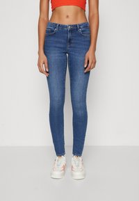 Blue skinny jeans with a mid-rise waist, featuring front pockets and a button closure. Paired with white and pink sneakers.