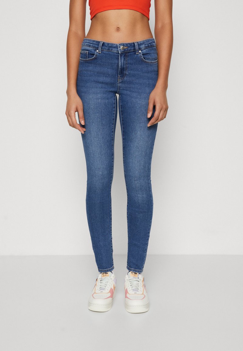 Blue skinny jeans with a mid-rise waist, featuring front pockets and a button closure. Paired with white and pink sneakers.