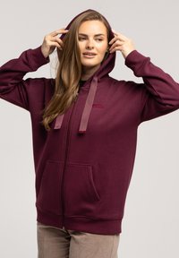 Bordowa bluza z kapturem na zamek, z regulowanym kapturem, kieszeniami z przodu oraz miękką teksturą. Zawiera metkę z logo na klatce piersiowej oraz prążkowane mankiety.